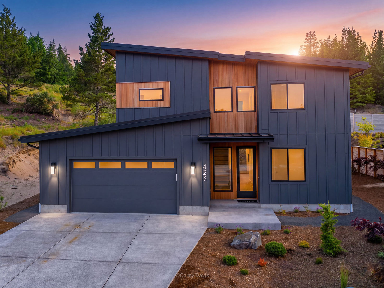 A modern two-storey house with dark cladding and a wide driveway, photographed at sunset.